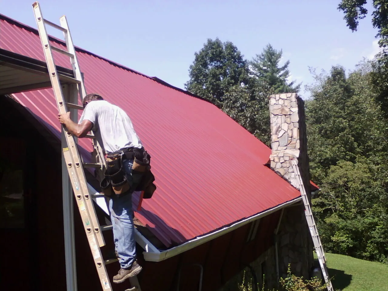 Roofer on a ladder beside a red metal roof and stone chimney — Soffit & Fascia Repair by Roofing Brothers in Victorville